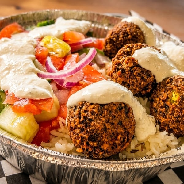 Falafel bowl with rice, salad, and white sauce in a foil container on a checkered paper liner on a checkered paper.